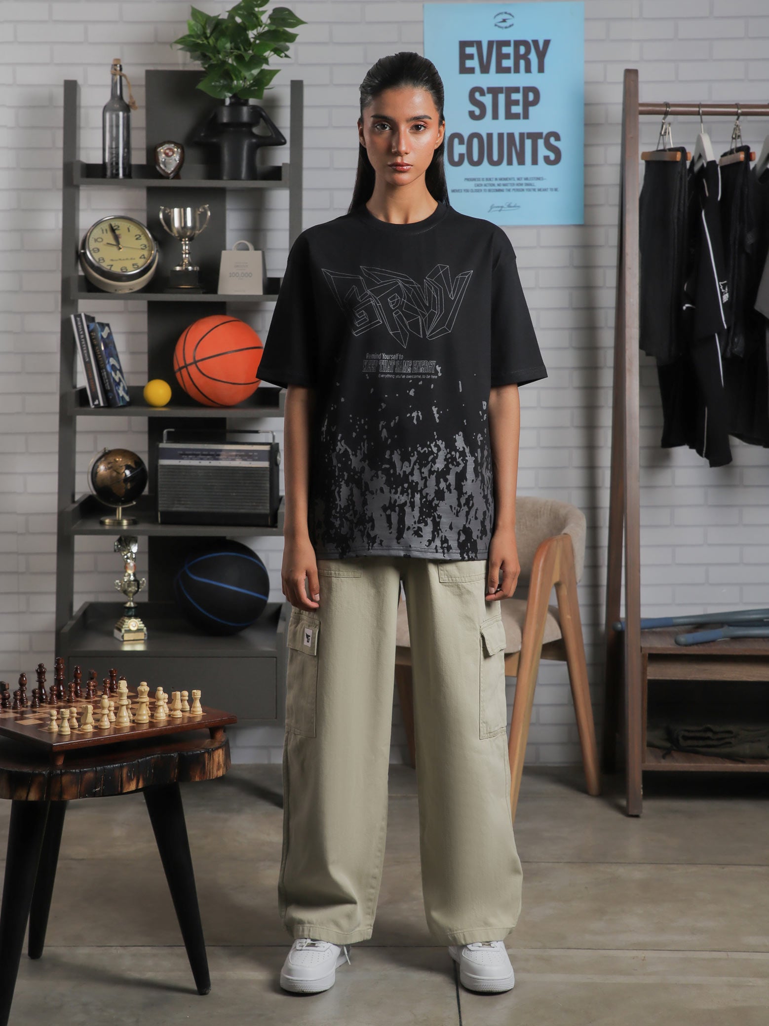Female model wearing “Keep That Same Energy” black oversized graphic t-shirt with khaki cargo pants, standing in studio with bookshelf and decor.
