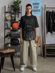 Female model wearing “Keep That Same Energy” black oversized graphic t-shirt with khaki cargo pants, standing in studio with bookshelf and decor.
