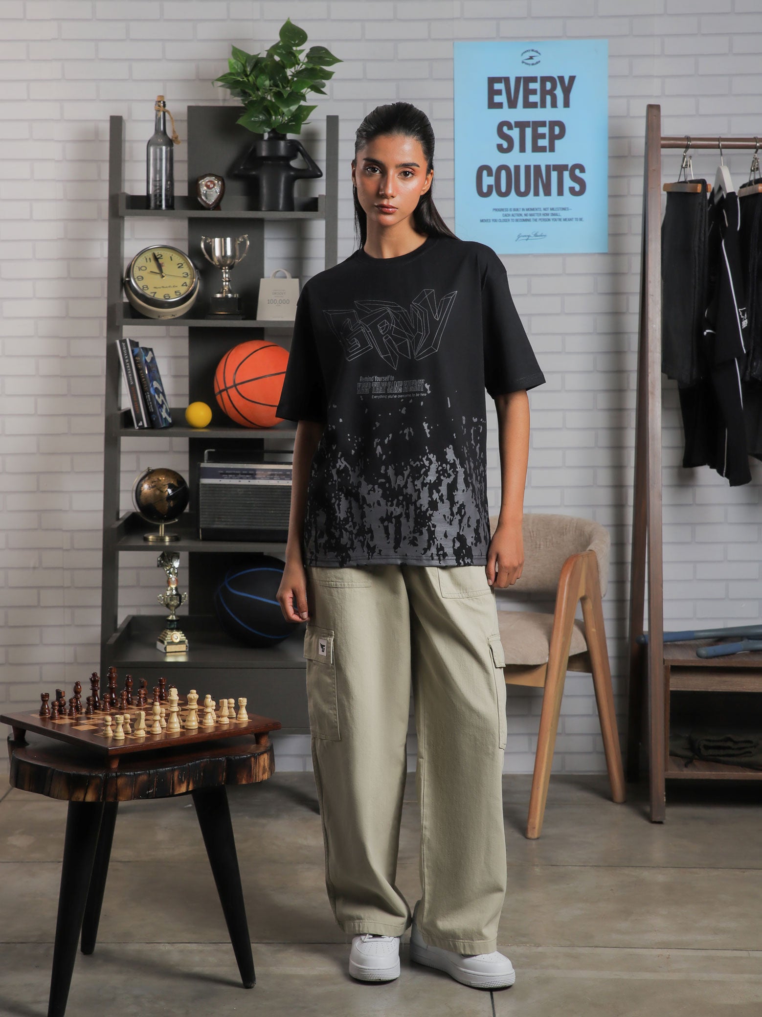 Female model wearing “Keep That Same Energy” black oversized graphic t-shirt with khaki cargo pants, standing in studio with bookshelf and decor.