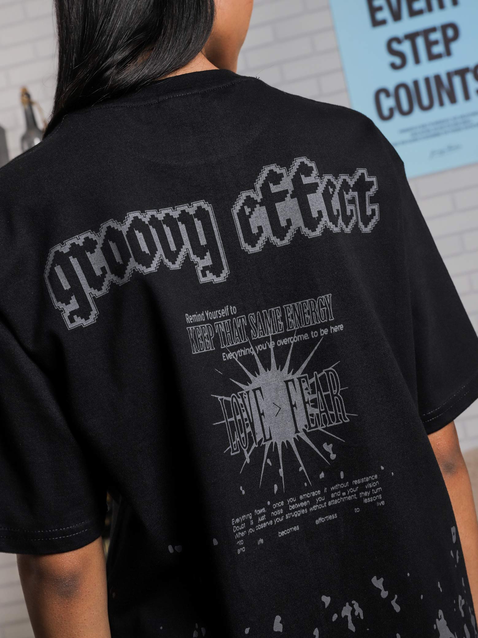 Back view of a black graphic t-shirt with pixel-style "groovy effect" text and an inspirational quote reading "Keep That Same Energy" and "Love > Fear" in a bold typographic layout, worn by a woman indoors with a poster in the background.