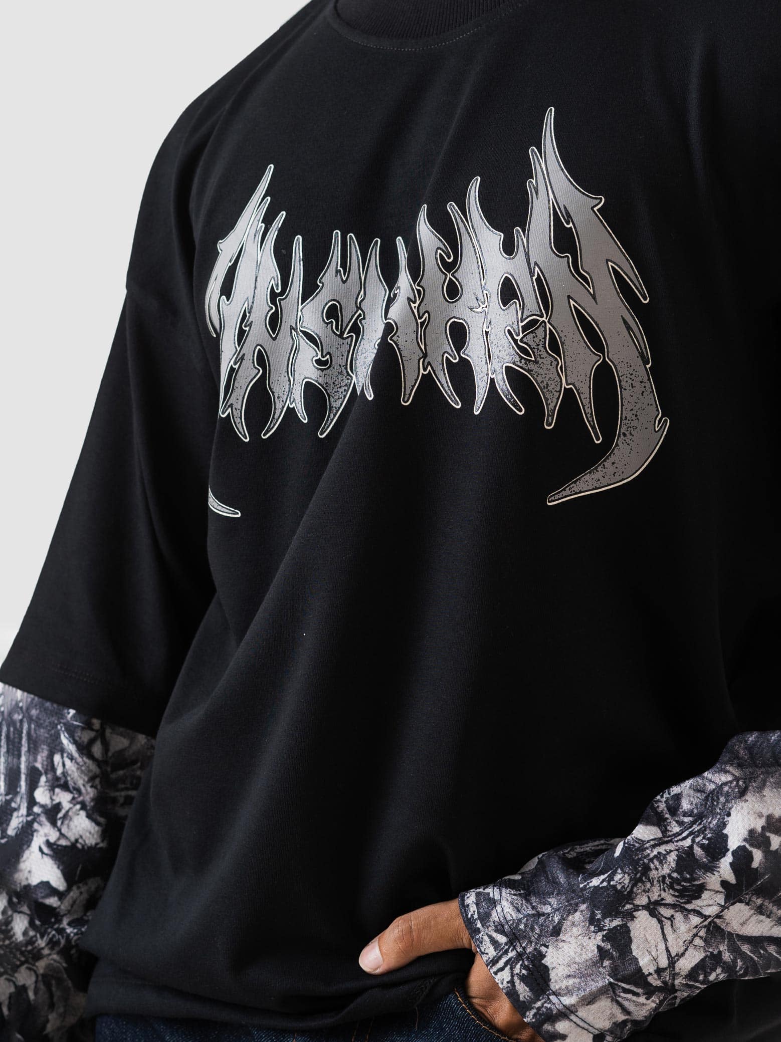 A close-up of a person wearing a black oversized graphic t-shirt with a metallic silver 'The Ashes' logo across the chest, layered over a black and white patterned long-sleeve shirt.